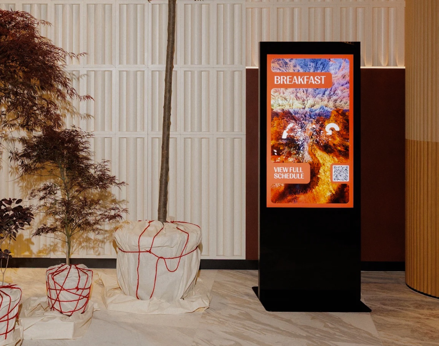 Sculptural tree and red wire-frame installations in a sunlit lobby beside a digital poster
